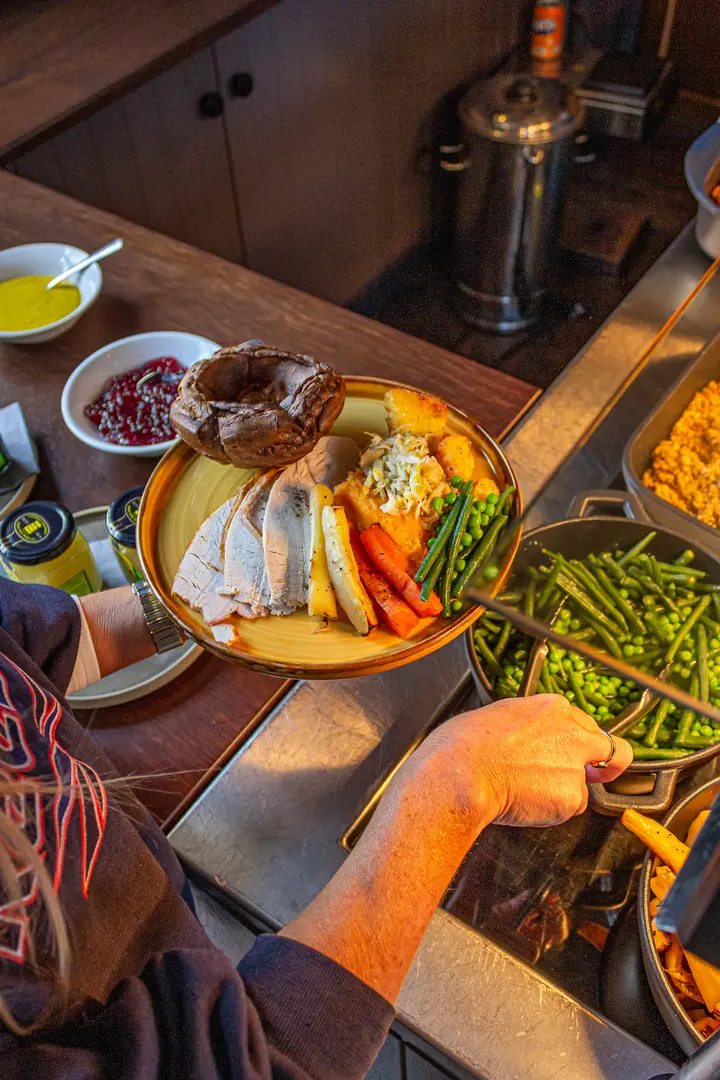 A carvery plate being served: sliced roast turkey, a Yorkshire pudding, roast potatoes, carrots, green beans and peas, with mustard and cranberry on the side