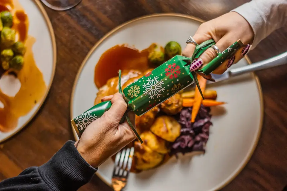 Two people pulling a green Christmas cracker over a plate of turkey, roast potatoes, carrots and red cabbage